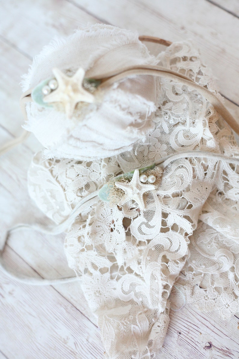 May include: Two white lace headbands with starfish and pearl accents. The headbands are on a white wooden background.