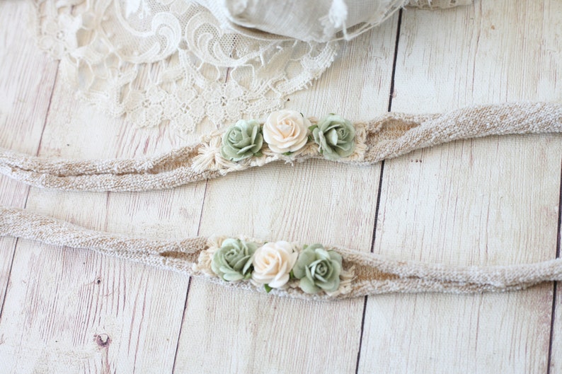 May include: Two beige fabric headbands with green and cream fabric flowers attached. The headbands are lying on a white wooden surface.
