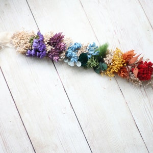 Puede incluir: Una guirnalda de flores secas de colores arco&iacute;ris con una cinta blanca. La guirnalda presenta flores moradas, azules, verdes, amarillas, naranjas y rojas.