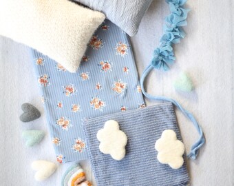 May include: An assortment of soft, pastel-colored items, including pillows, fabric, and decorative accents. The image features a light blue pillow with a textured knit design, a white pillow, and a blue floral fabric. Small felt hearts and cloud-shaped decorations are also present.