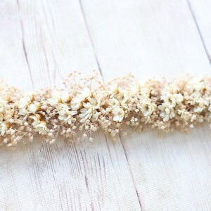 May include: A garland of dried white flowers and tan stems on a white wood background.