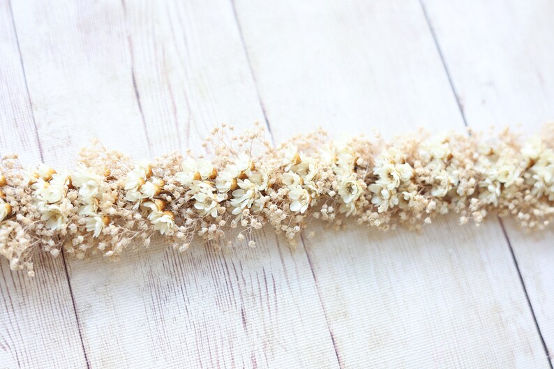 May include: A garland of dried white flowers and tan stems on a white wood background.