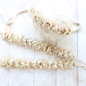 May include: Two white flower crowns, one is a headband and the other is a longer garland, both made with dried white flowers and a beige ribbon.
