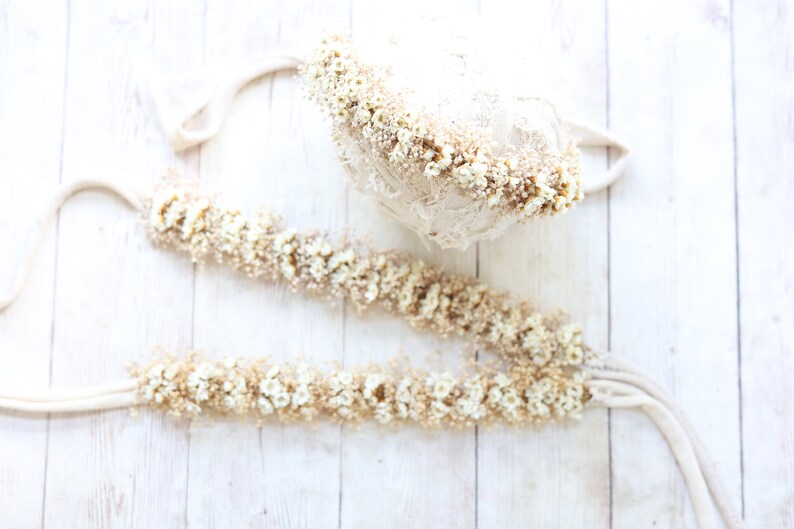 May include: Two white flower crowns, one is a headband and the other is a longer garland, both made with dried white flowers and a beige ribbon.