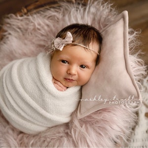 May include: A newborn baby girl is swaddled in a white knitted blanket and lying on a pink fluffy surface. She is wearing a pink bow headband and looking directly at the camera.