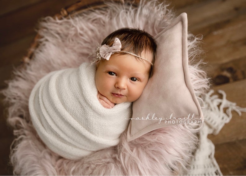 May include: A newborn baby girl is swaddled in a white knitted blanket and lying on a pink fluffy surface. She is wearing a pink bow headband and looking directly at the camera.