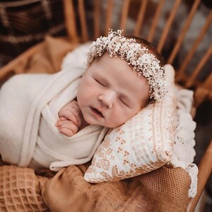 May include: A newborn baby girl wearing a white flower crown sleeps peacefully on a white pillow with a floral pattern. The baby is wrapped in a white blanket and is lying on a brown woven chair.