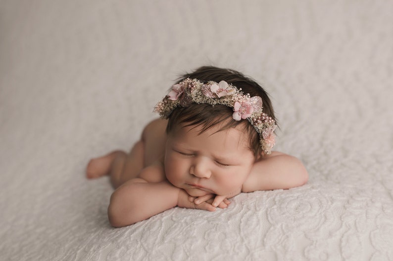 May include: A newborn baby wearing a pink flower crown is sleeping on a white textured blanket. The baby is lying on their stomach with their arms crossed under their chin.