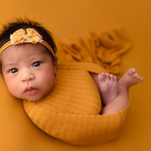 May include: A newborn baby wrapped in a mustard yellow knitted blanket, with a matching headband featuring a flower. The baby is positioned on a solid mustard yellow background, with the baby's feet visible.