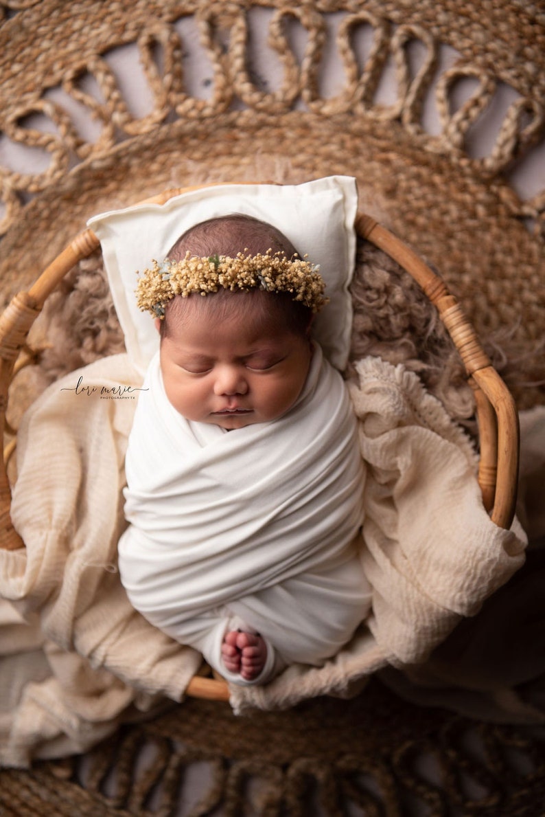 May include: A newborn baby wearing a white swaddle and a flower crown is sleeping in a wicker basket. The baby is surrounded by a soft, beige blanket.