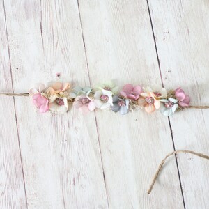 Puede incluir: Una diadema floral con flores de tela en tonos pastel, rosa, melocot&oacute;n y azul, ensartadas en hilo natural. Las flores est&aacute;n agrupadas, creando un accesorio delicado y femenino para el cabello.