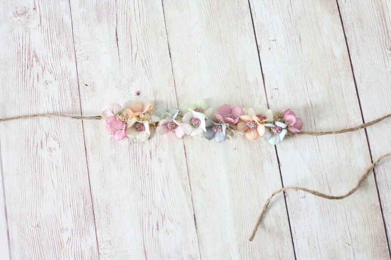 Puede incluir: Una diadema floral con flores de tela en tonos pastel, rosa, melocot&oacute;n y azul, ensartadas en hilo natural. Las flores est&aacute;n agrupadas, creando un accesorio delicado y femenino para el cabello.