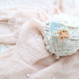 May include: A white headband with a light blue felt heart and a starfish charm. The headband is on a pink fabric background.