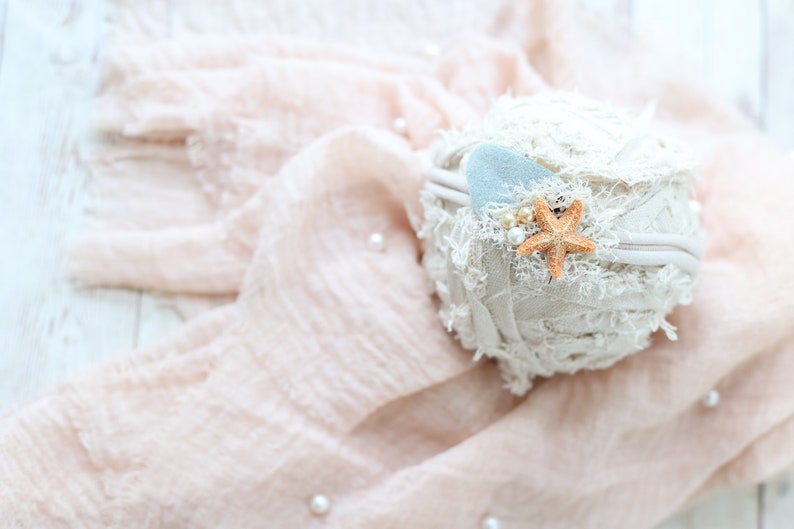 May include: A white headband with a light blue felt heart and a starfish charm. The headband is on a pink fabric background.