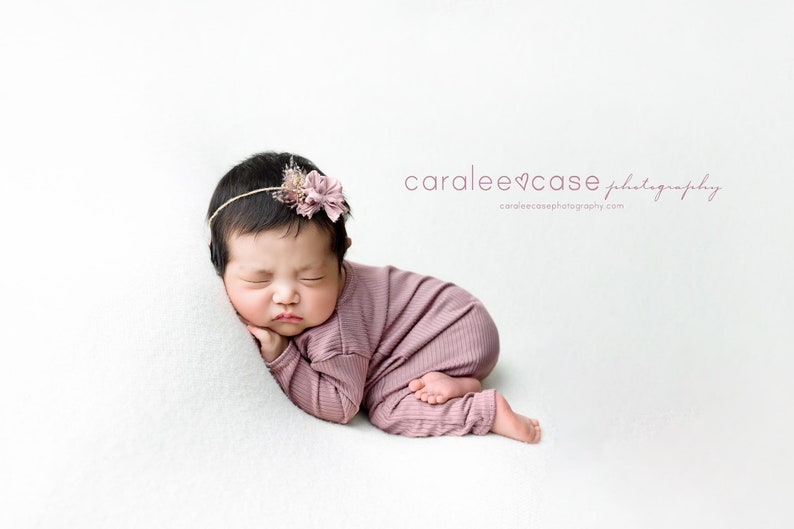 May include: A newborn baby girl is sleeping on a white background. She is wearing a pink knitted onesie and a headband with a pink flower. The text "caralee&hearts;case photography" is visible in the upper right corner of the image.