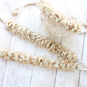 May include: Two white fabric garlands decorated with dried white flowers and greenery. The garlands are laid out on a white wooden surface.