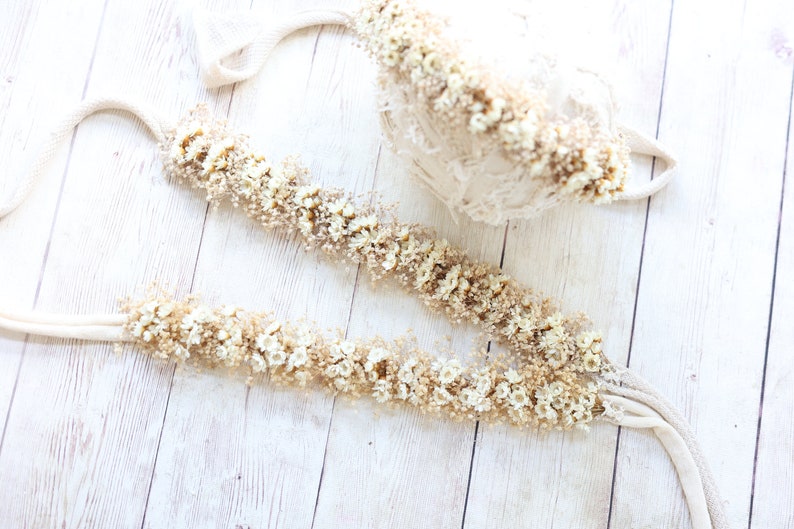 May include: Two white fabric garlands decorated with dried white flowers and greenery. The garlands are laid out on a white wooden surface.