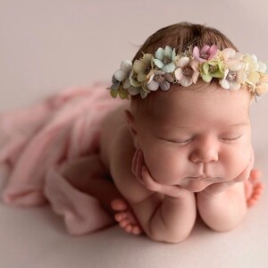 May include: A baby wearing a floral crown, lying with its eyes closed. The baby is resting its head on its hands and is wrapped in a soft pink fabric. The crown features pastel-coloured flowers, creating a gentle image.