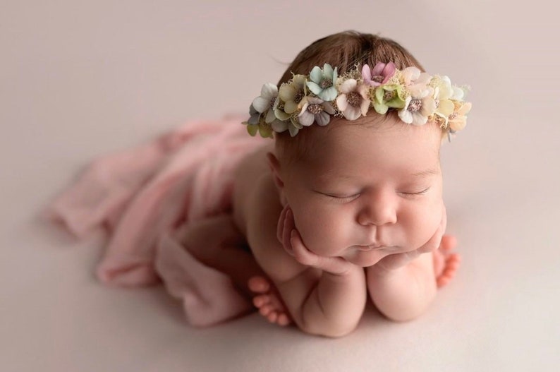 May include: A baby wearing a floral crown, lying with its eyes closed. The baby is resting its head on its hands and is wrapped in a soft pink fabric. The crown features pastel-coloured flowers, creating a gentle image.