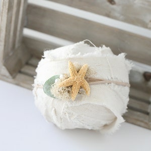 May include: A white fabric headband with a starfish and green leaf embellishment. The headband is rolled up and sitting on a wooden crate.