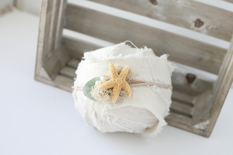 May include: A white fabric headband with a starfish and green leaf embellishment. The headband is rolled up and sitting on a wooden crate.