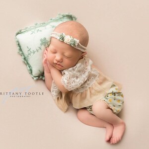 May include: A newborn baby girl sleeping on a green pillow with a floral design. She is wearing a beige lace top and green floral print pants with a white flower headband.