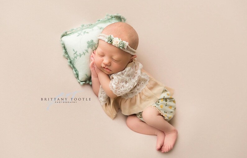 May include: A newborn baby girl sleeping on a green pillow with a floral design. She is wearing a beige lace top and green floral print pants with a white flower headband.
