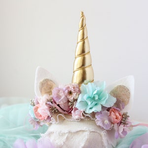 May include: A white unicorn headband with a gold spiral horn, pink, purple, and blue flowers, and white felt ears with gold glitter accents.