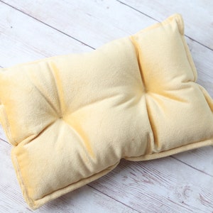 Pode incluir: Uma pequena almofada retangular amarelo claro com um design tufado. A almofada tem uma textura macia e felpuda e é provavelmente usada para fins decorativos ou como uma pequena almofada. A almofada é de cor sólida e tem um design simples e clássico.