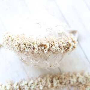 May include: A white and beige newborn baby photo prop with a delicate lace design and a band of dried white flowers around the top.