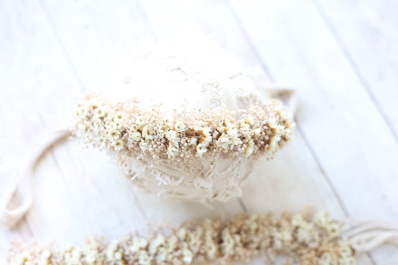 May include: A white and beige newborn baby photo prop with a delicate lace design and a band of dried white flowers around the top.