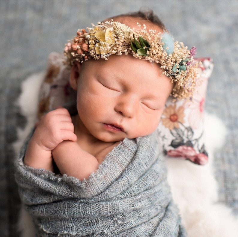 Puede incluir: Una beb&eacute; durmiendo con una corona de flores secas y envuelta en una manta de tela gris.
