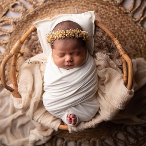 May include: A newborn baby wearing a white swaddle and a flower crown is sleeping in a wicker basket. The basket is on a woven rug.