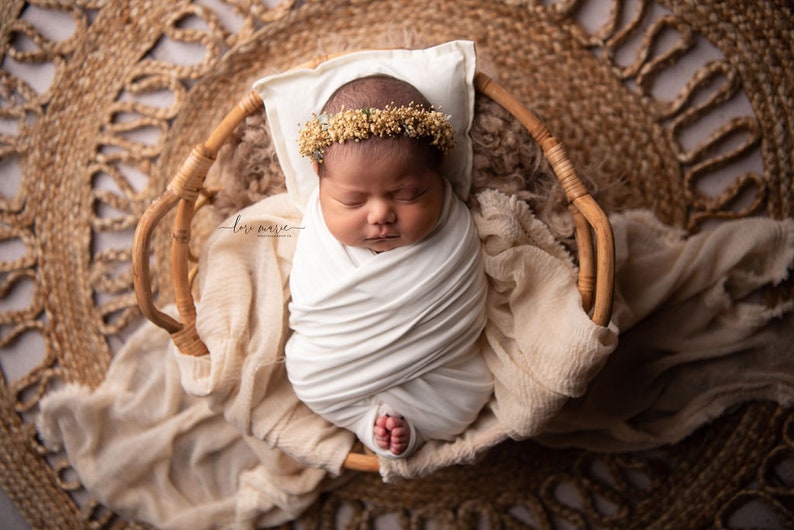 May include: A newborn baby wearing a white swaddle and a flower crown is sleeping in a wicker basket. The basket is on a woven rug.