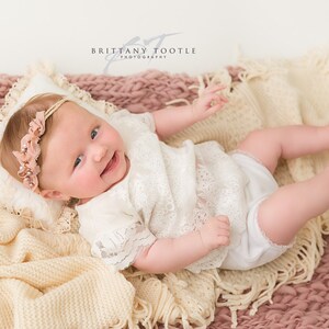 May include: A smiling baby wearing a white lace outfit and a floral headband, lying on a cream-colored pillow and blankets. The blankets are in shades of cream and pink. The baby is the focal point of the image.