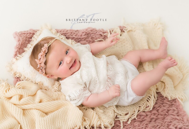 May include: A smiling baby wearing a white lace outfit and a floral headband, lying on a cream-colored pillow and blankets. The blankets are in shades of cream and pink. The baby is the focal point of the image.
