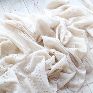 May include: A white crocheted blanket with a delicate openwork pattern. The blanket is made of soft, lightweight yarn and has a relaxed, drapey feel.