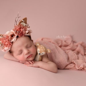 May include: A newborn baby girl wearing a pink and brown floral headband with antlers sleeps on a pink blanket with a small stuffed deer toy.