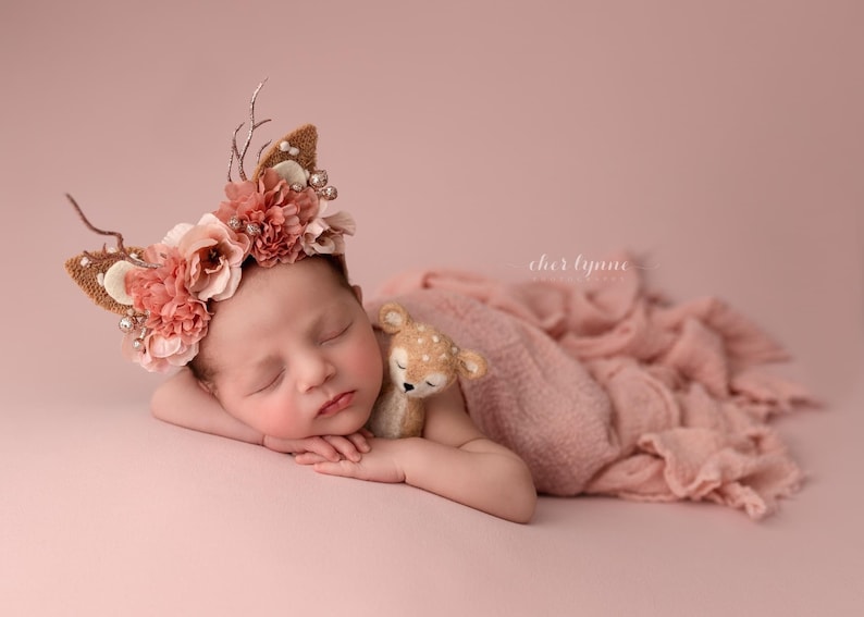 May include: A newborn baby girl wearing a pink and brown floral headband with antlers sleeps on a pink blanket with a small stuffed deer toy.