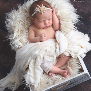 May include: A newborn baby girl sleeping in a white wooden box lined with a white fluffy blanket. The baby is wearing a white headband with a starfish design. The box has a rope handle.