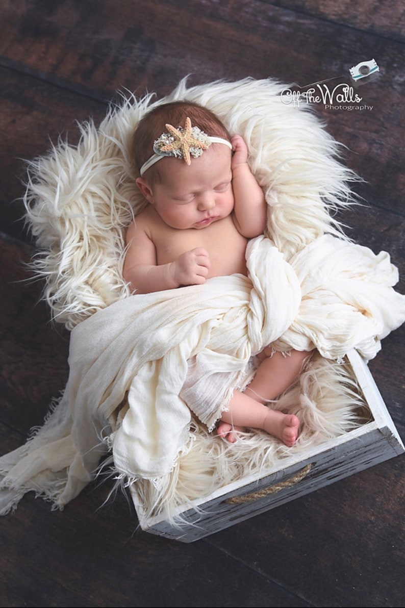 May include: A newborn baby girl sleeping in a white wooden box lined with a white fluffy blanket. The baby is wearing a white headband with a starfish design. The box has a rope handle.