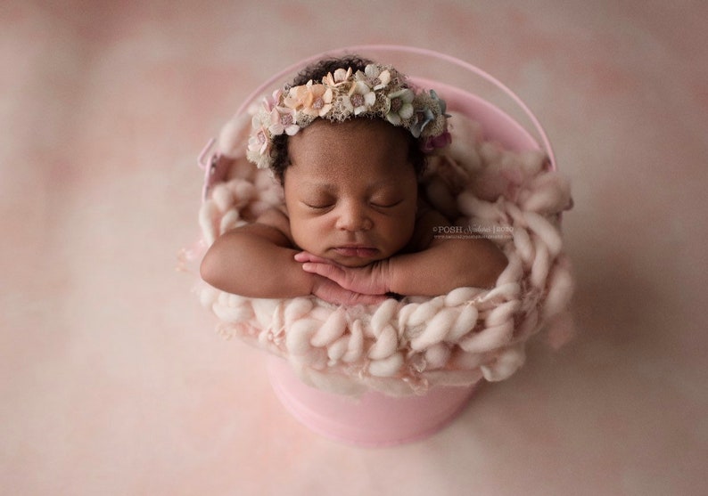 Puede incluir: Un beb&eacute; reci&eacute;n nacido con una corona de flores descansa pl&aacute;cidamente en un cubo rosa lleno de lana suave de color rosa claro. Los brazos del beb&eacute; est&aacute;n cruzados y los ojos cerrados. El fondo es de color rosa claro.