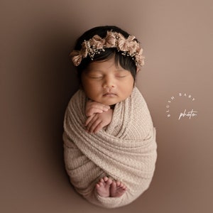 May include: A newborn baby wrapped in a beige knitted blanket, sleeping with a flower crown on their head. The baby is lying on a brown background with the text "Blush Baby Photo" in a circle.