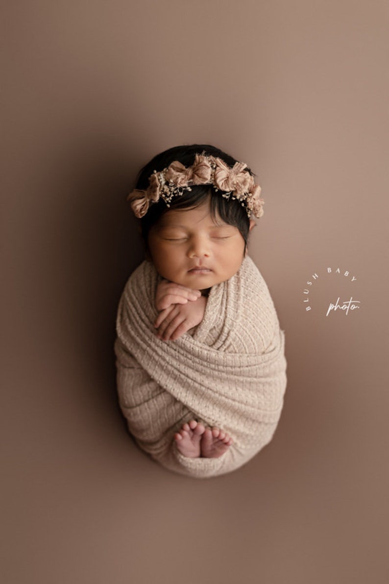 May include: A newborn baby wrapped in a beige knitted blanket, sleeping with a flower crown on their head. The baby is lying on a brown background with the text "Blush Baby Photo" in a circle.