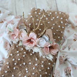 May include: A brown knitted baby blanket with white polka dots and a floral headband with gold antlers. The headband has pink and white flowers and a brown felt base.
