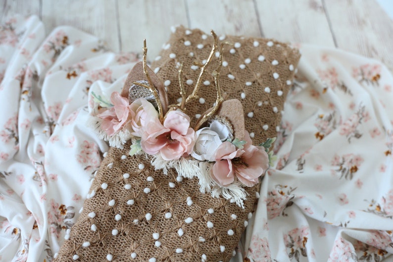 May include: A brown knitted baby blanket with white polka dots and a floral headband with gold antlers. The headband has pink and white flowers and a brown felt base.