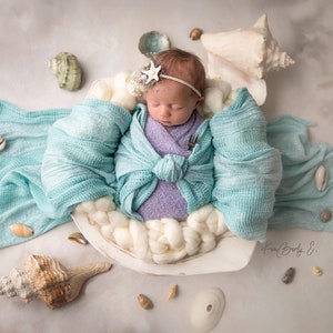 May include: A newborn baby is sleeping in a white seashell with a light blue blanket. The baby is wearing a purple swaddle and a white star headband. The seashell is surrounded by small seashells.