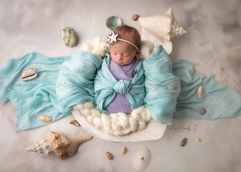 May include: A newborn baby is sleeping in a white seashell with a light blue blanket. The baby is wearing a purple swaddle and a white star headband. The seashell is surrounded by small seashells.