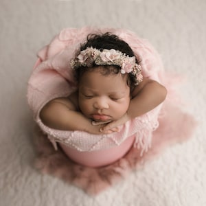 May include: A newborn baby wearing a pink flower crown is sleeping in a pink bucket. The baby is wrapped in a pink blanket.