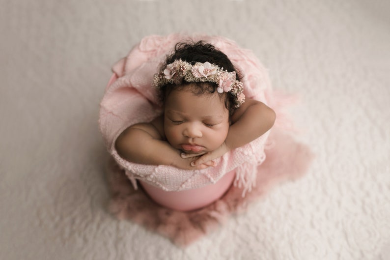 May include: A newborn baby wearing a pink flower crown is sleeping in a pink bucket. The baby is wrapped in a pink blanket.